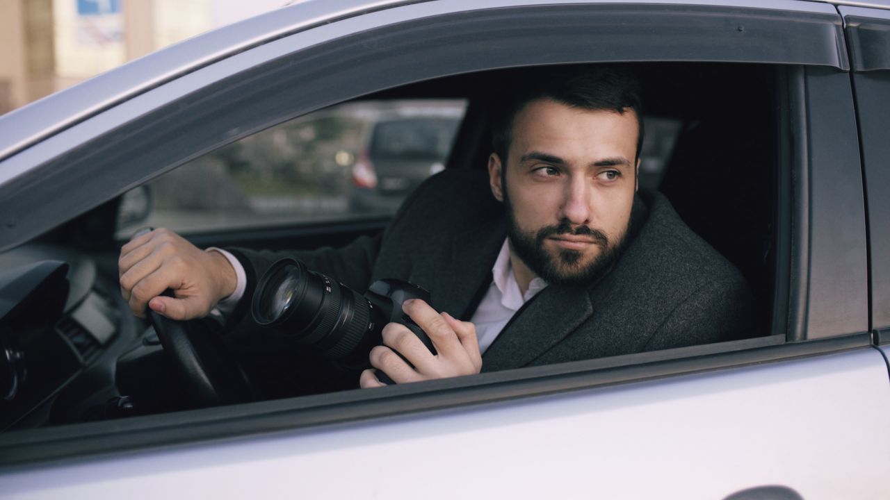 Home A bearded man in a car holding a camera, ready to take photos outdoors.