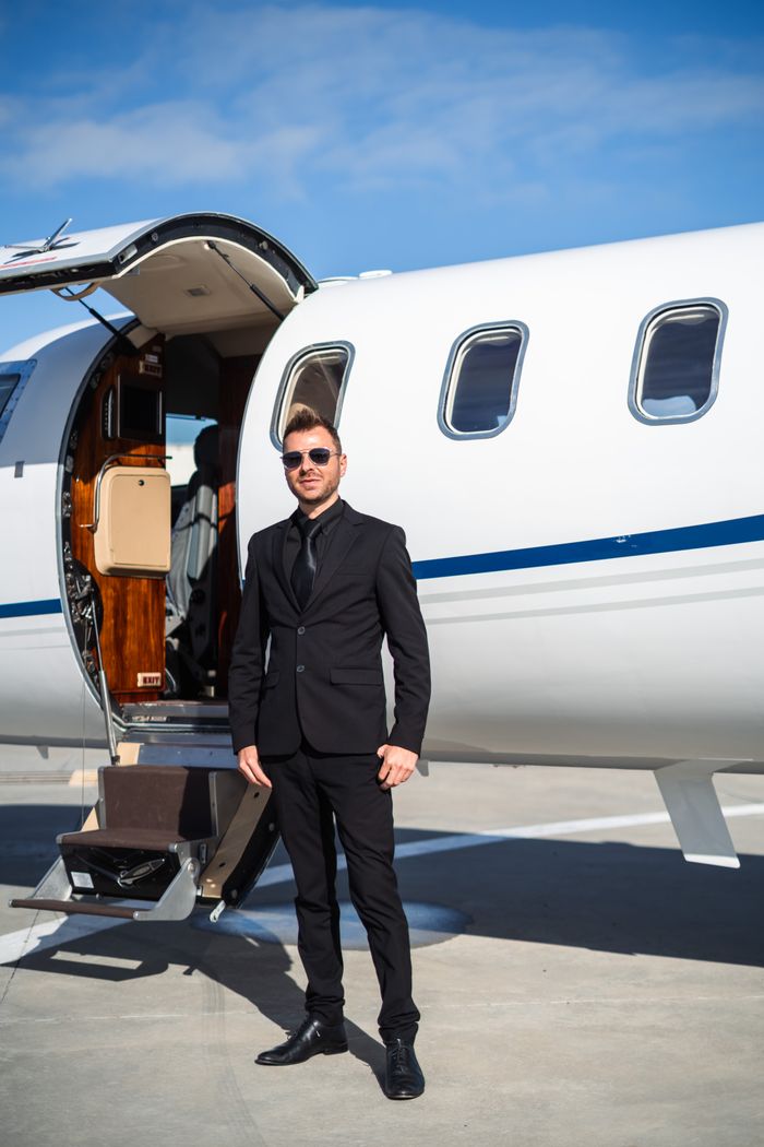 Home Stylish man in black suit poses confidently beside a private jet under clear blue skies.