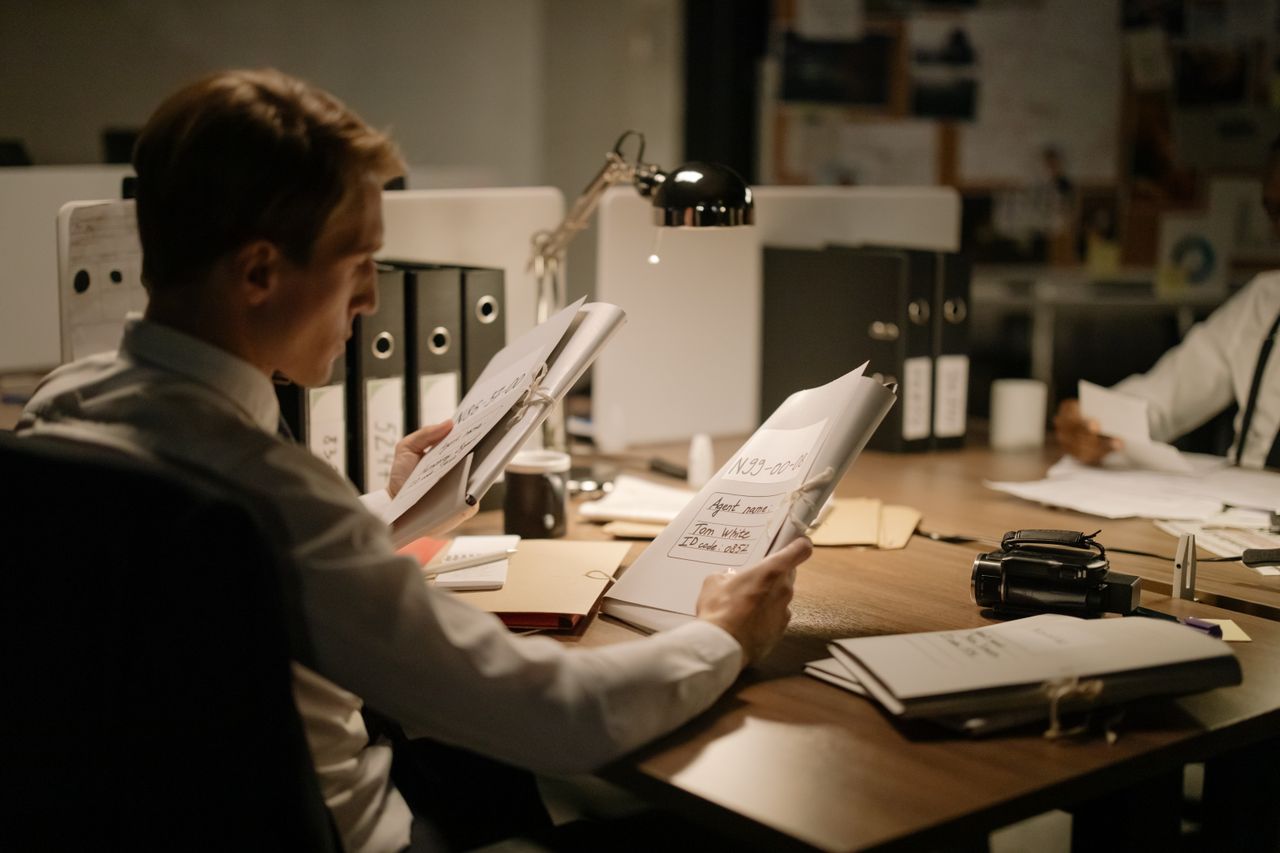 Two detectives reviewing documents in a dimly lit office setting, focused and professional.
